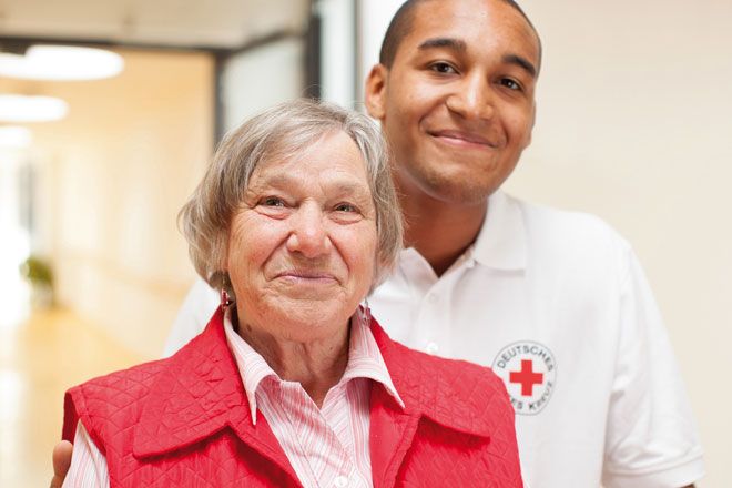 Foto: Ein junger Mann im Freiwilligendienst begleitet eine ältere Dame durch die Gänge einer stationären Einrichtung. Die Dame scheint sich über ihre Begleitung sichtlich zu freuen.